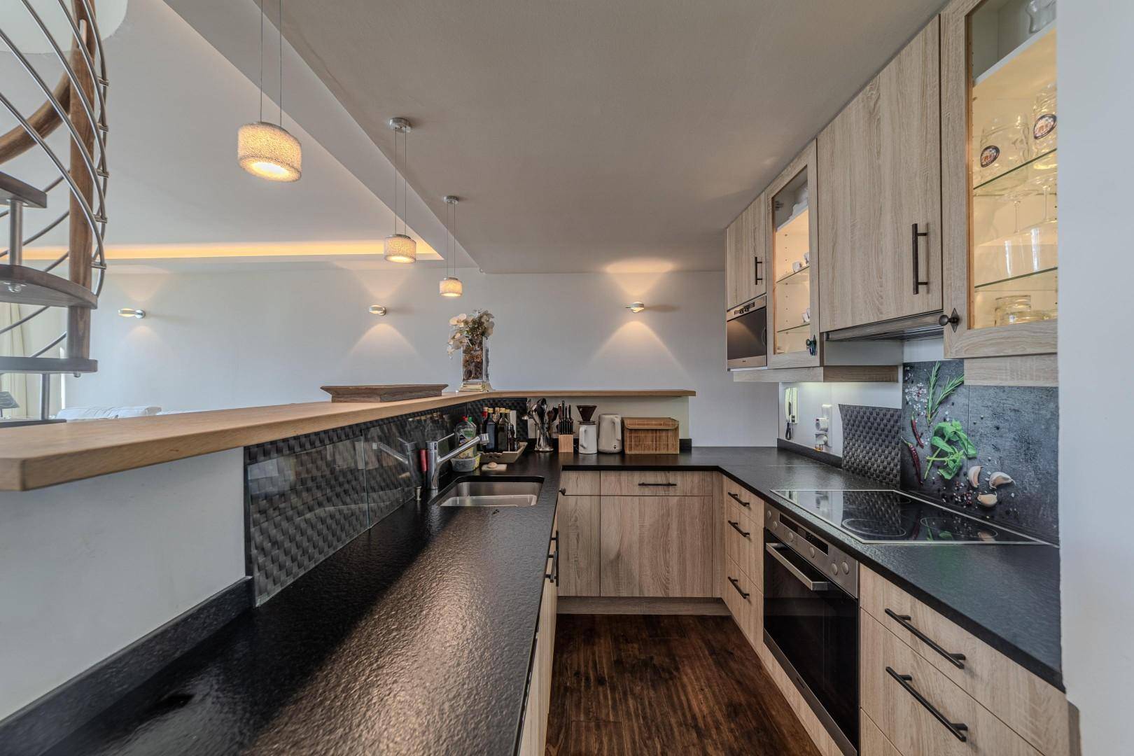 Moderne Küche mit Holzfronten, dunkler Arbeitsplatte und integrierten Geräten, offen zum Wohnbereich.