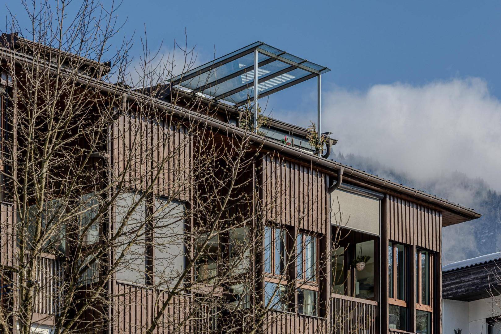 Detailansicht einer modernen Holzfassade mit großen Fenstern und einer Glasüberdachung auf dem Dach.
