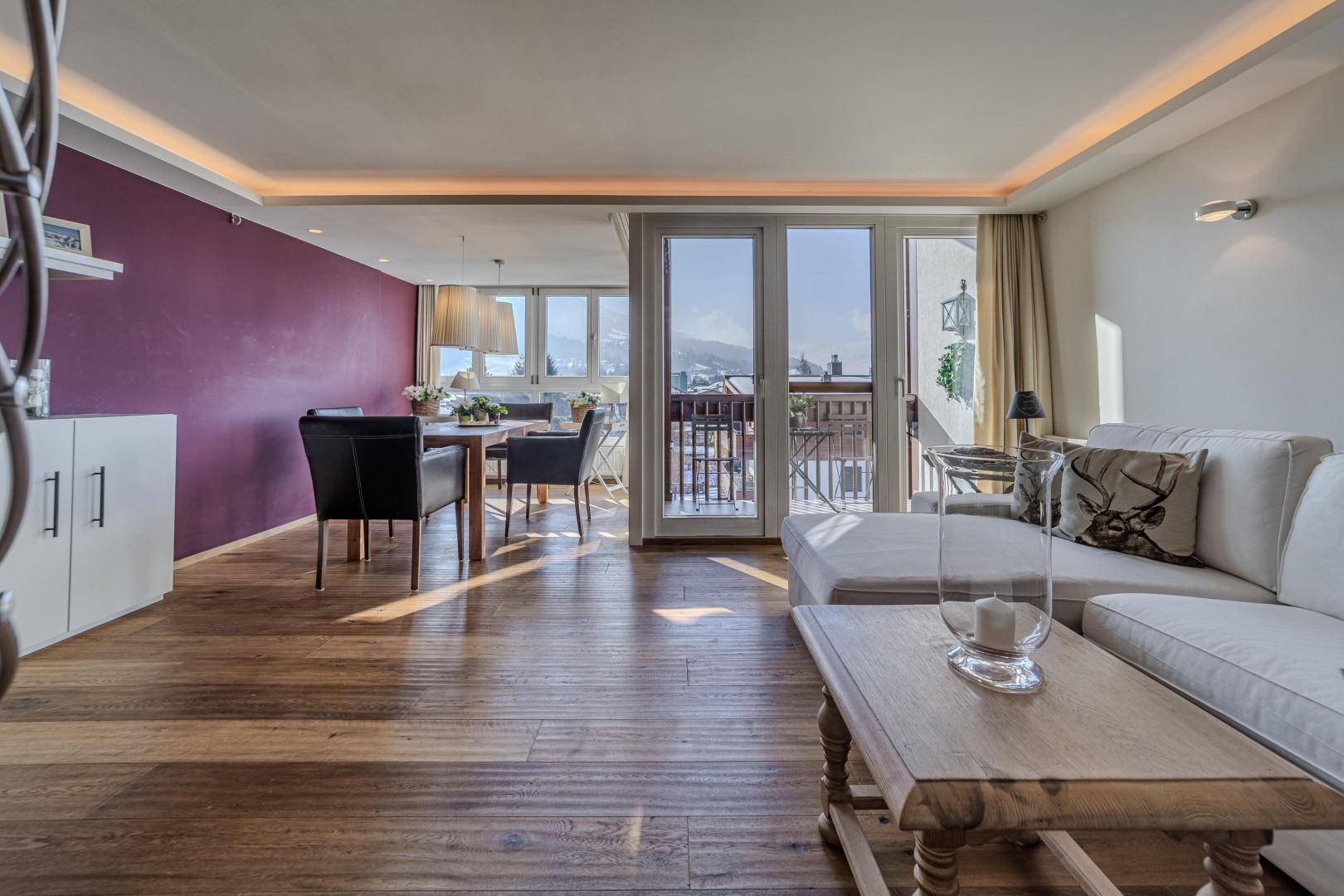 Offener Wohn- und Essbereich mit Holzboden, farbiger Wand und Zugang zu einem sonnigen Balkon.