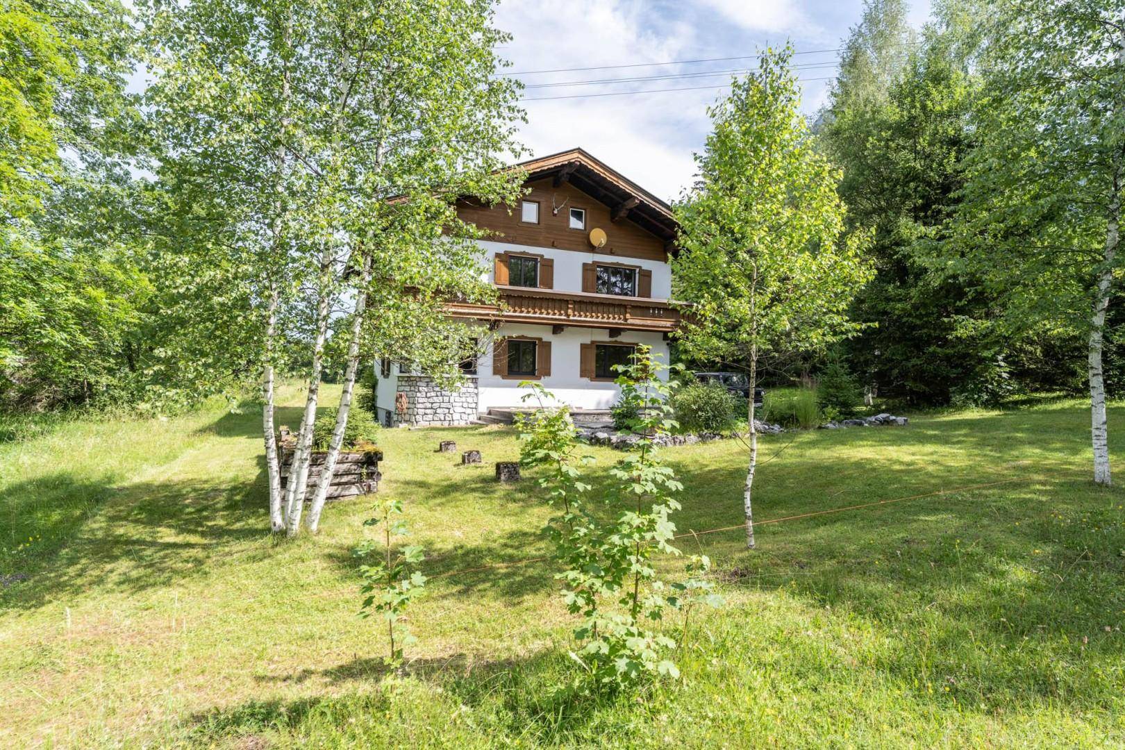 Traditionelles Haus auf einem grünen Hügel, umgeben von Birken und anderen Bäumen in ländlicher Idylle.