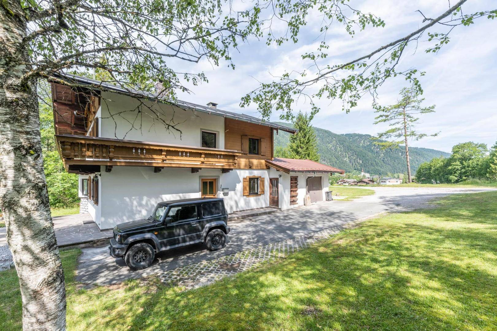 Charmantes Haus mit Holzbalkon und gepflasterter Auffahrt, umgeben von üppigem Grün und Bergen.