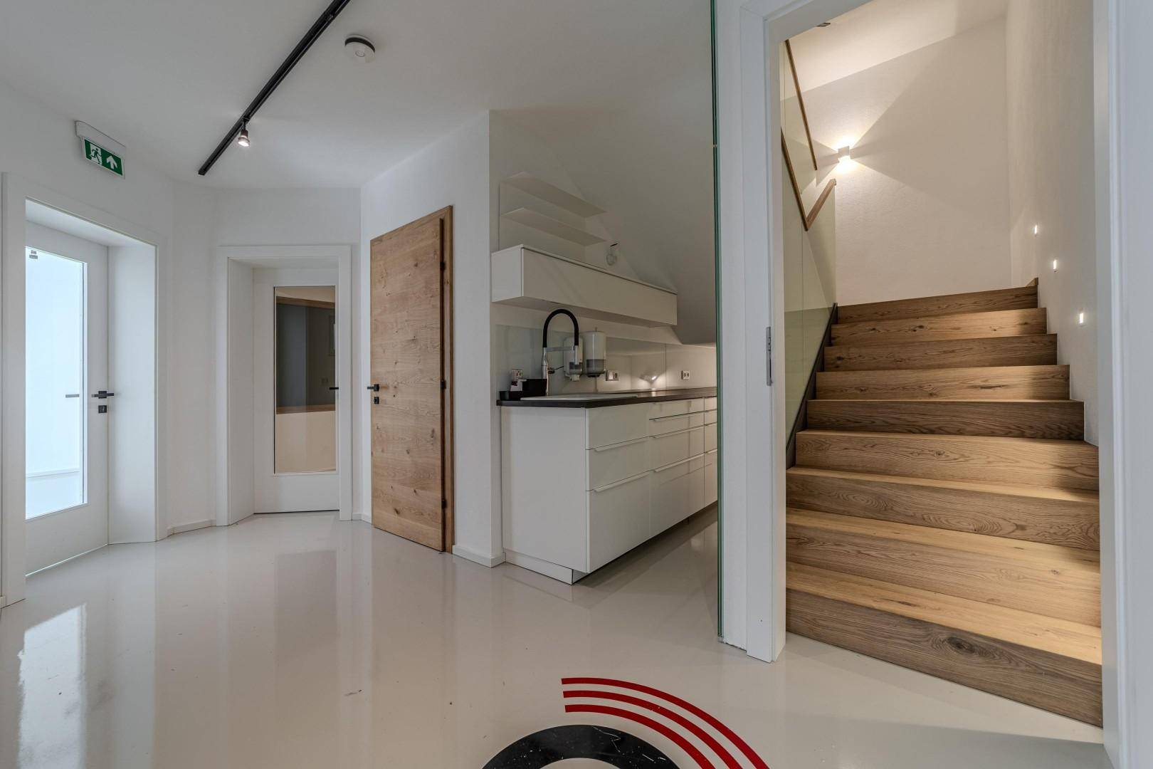 Heller Flur mit kleiner Küchenzeile und Holztreppe mit Glasgeländer, modern gestaltet.