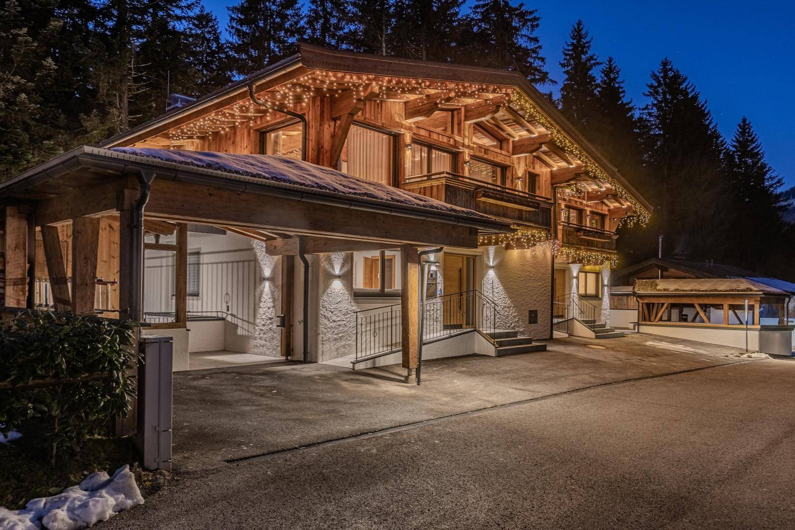 Chalet bei Nacht mit festlicher Beleuchtung und integriertem Carport, umgeben von verschneiten Bäumen.