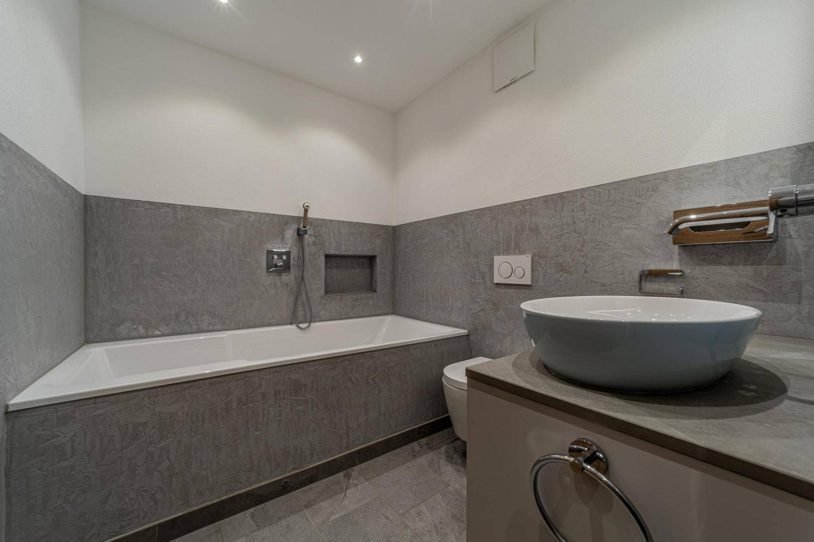 Modernes Badezimmer mit Badewanne, Aufsatzwaschbecken und grauen Fliesen an Wänden und Boden.