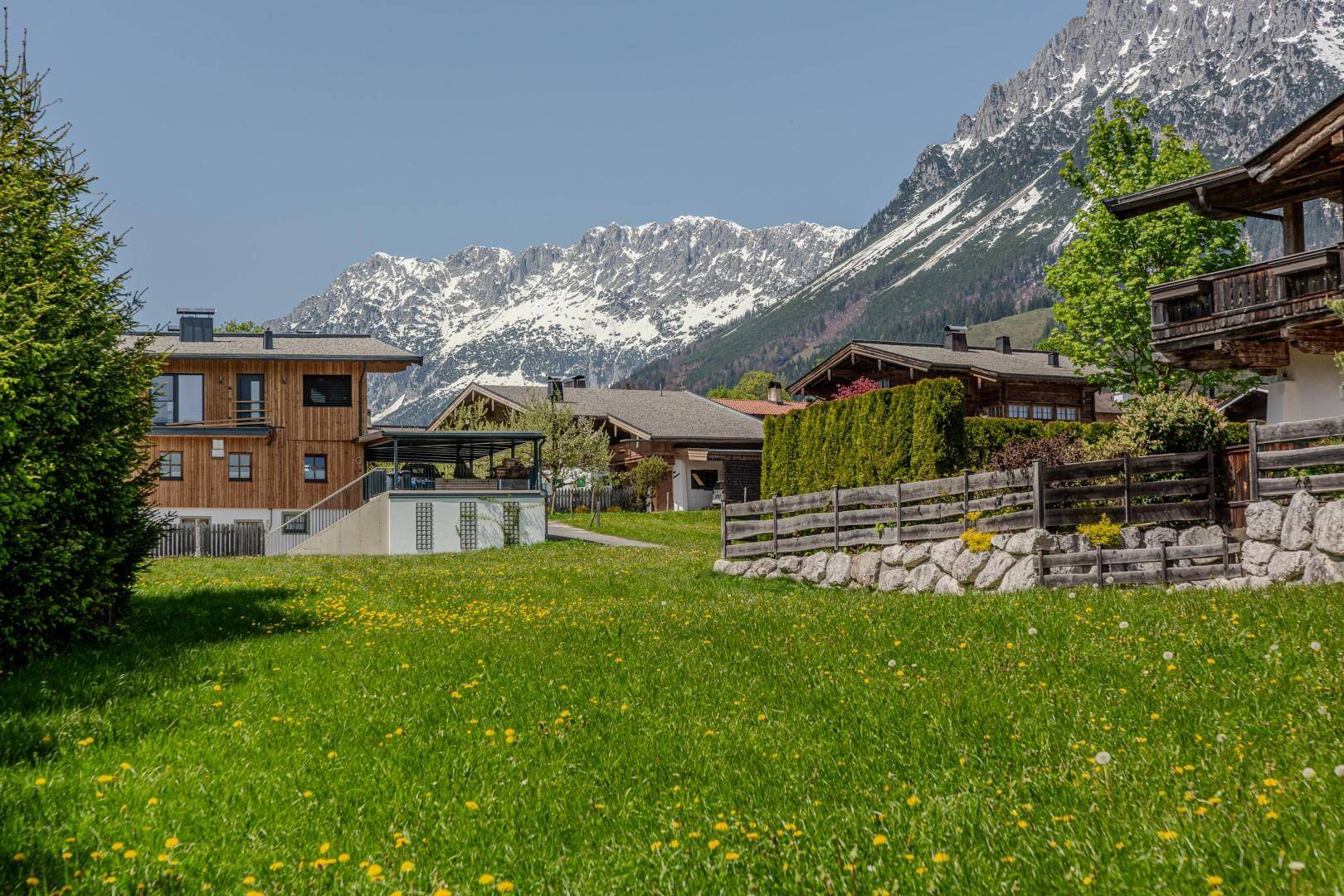 Wohngebiet mit mehreren Holzhäusern und grünen Wiesen vor einer beeindruckenden Bergkulisse.
