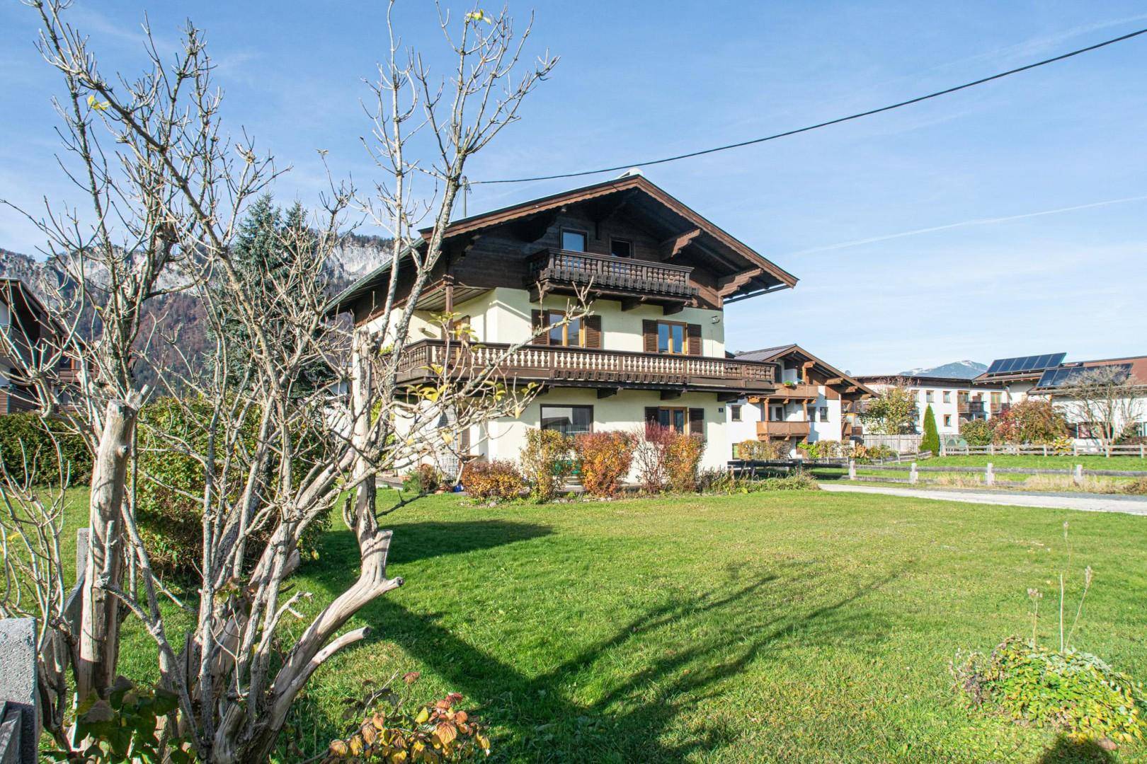 Vorderansicht des traditionellen Hauses mit Balkonen, umgeben von einem grünen Garten und Bergen.