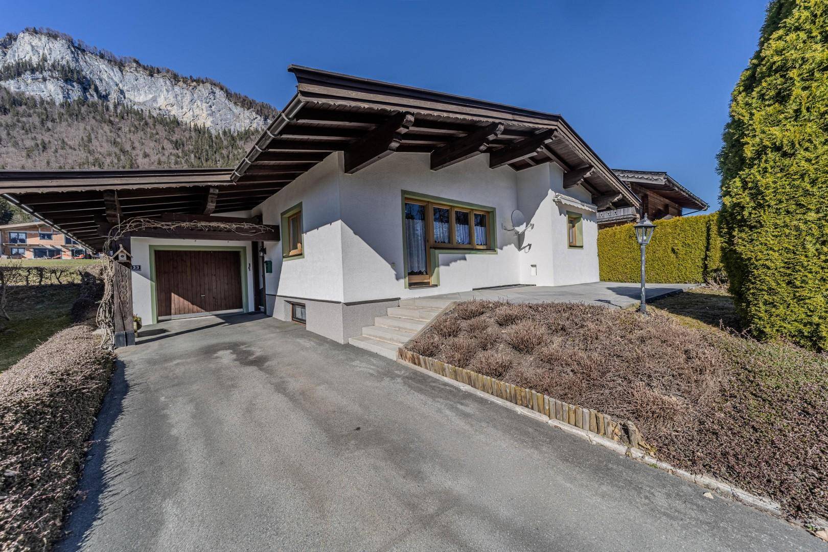 Ansicht des Hauses mit Zufahrt, Carport und Garage vor einer beeindruckenden Bergkulisse.