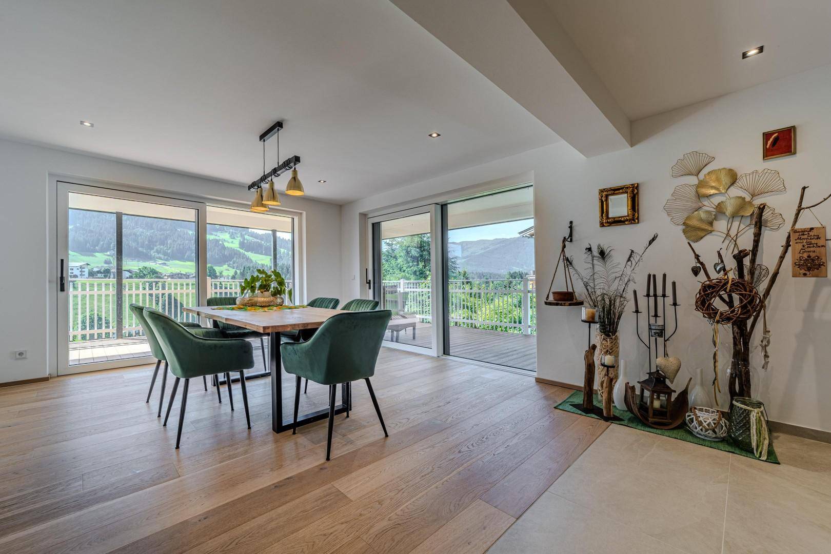 Heller Essbereich mit Holztisch und grünen Stühlen, Zugang zur Terrasse und Blick auf die grüne Berglandschaft.