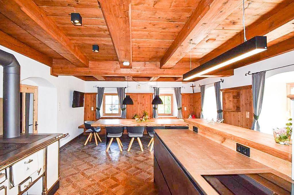 Rustikaler Essbereich mit langem Holztisch, Holzbalkendecke und traditionellem Ofen im Hintergrund.