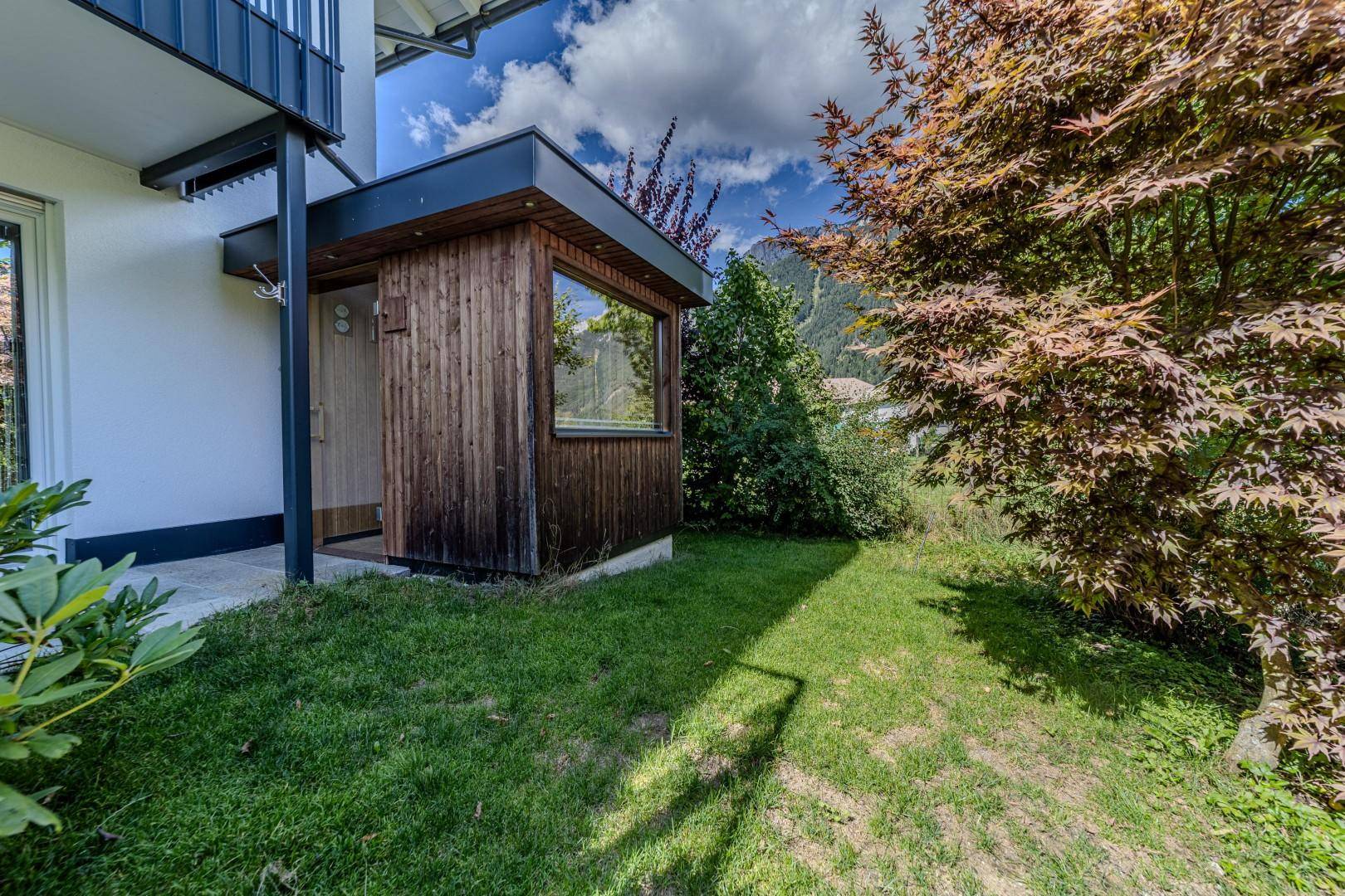 Sonniger Gartenbereich mit Rasen, Bäumen und einem kleinen Holzhaus im Hintergrund.