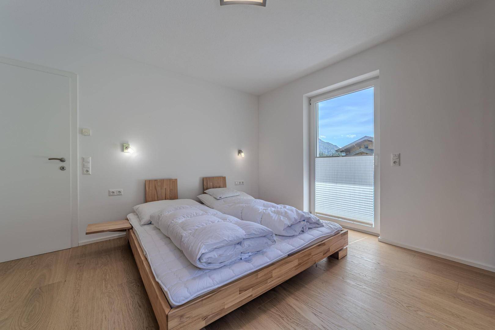 Helles Schlafzimmer mit Holzbett, weißen Wänden und Fenster mit Bergblick.