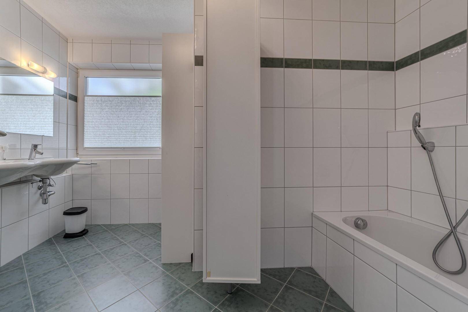 Helles Badezimmer mit Badewanne, Fenster und grünen Bodenfliesen.