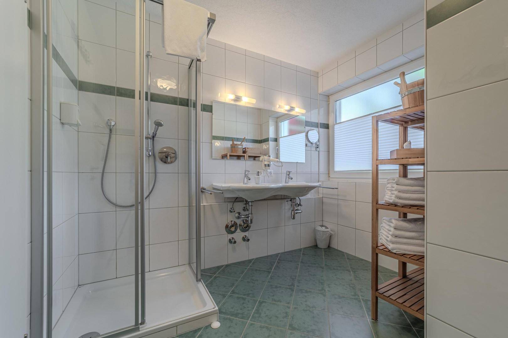 Helles Badezimmer mit Duschkabine, Doppelwaschbecken und Fenster, gefliest in Weiß und Grün.
