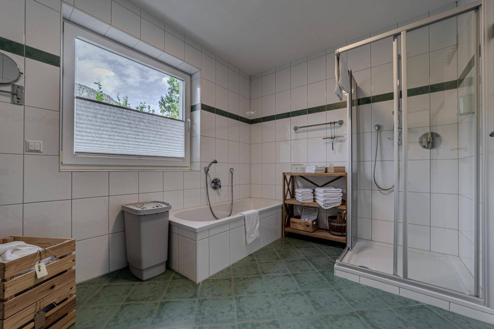 Helles Badezimmer mit Badewanne, Duschkabine und Fenster, gefliest in Weiß und Grün.