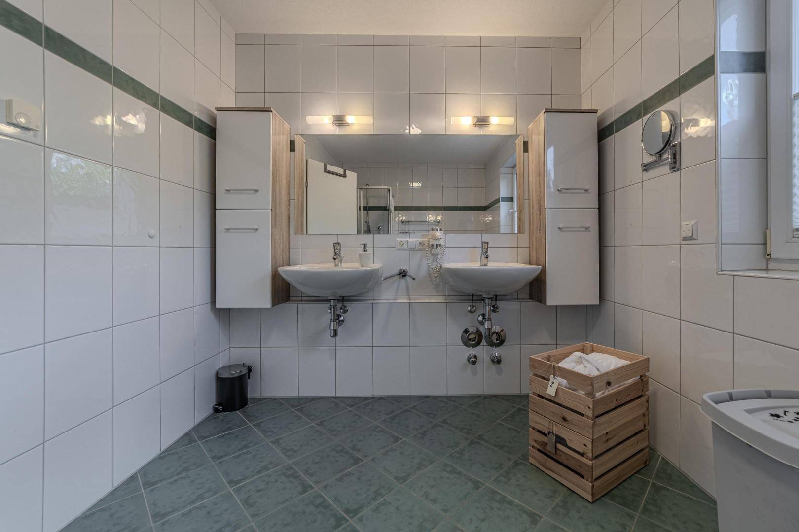 Geräumiges Badezimmer mit Doppelwaschbecken, großen weißen Fliesen und einem Fenster.