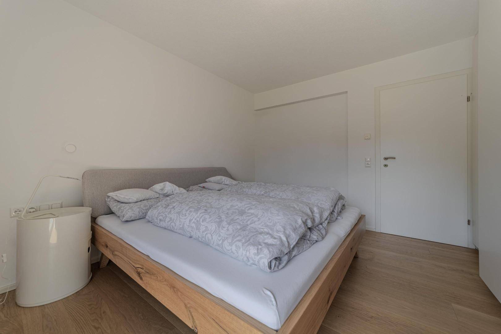 Ruhiges Schlafzimmer mit Holzbett, weißen Wänden und schlichtem Design.