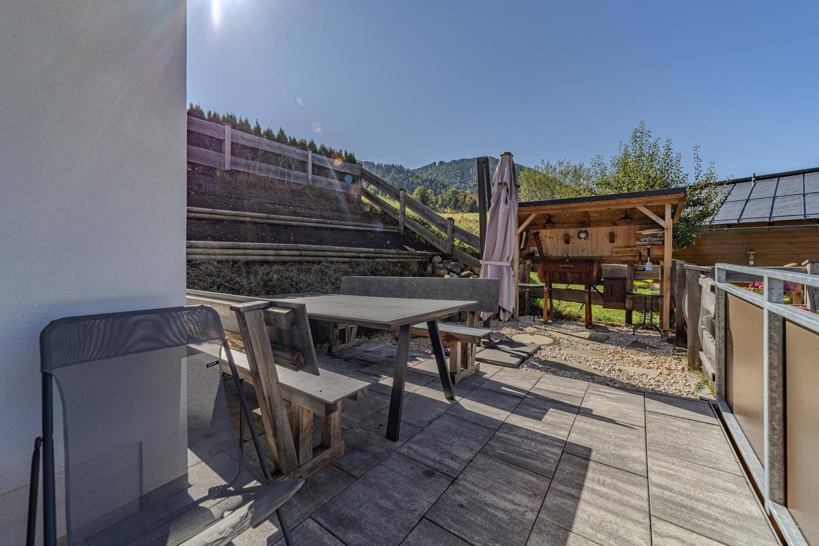 Sonniger Terrassenbereich mit Esstisch und integrierter Grillstation, umgeben von Natur.