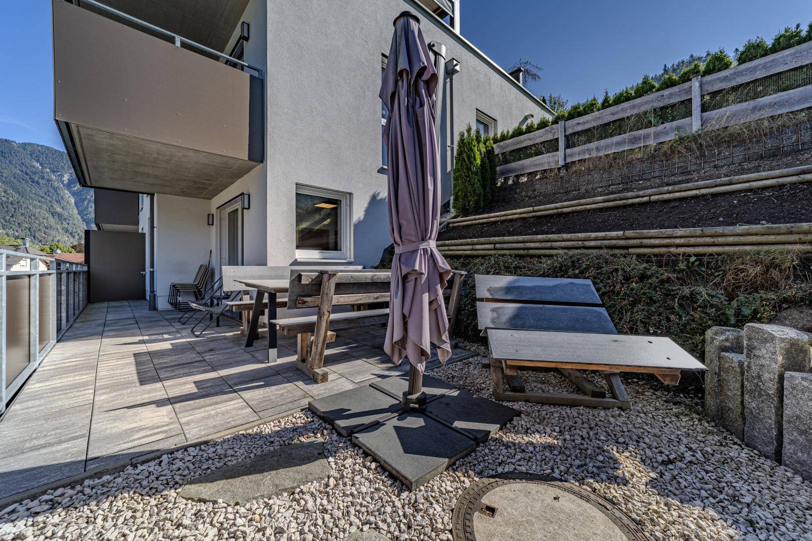 Geräumige Terrasse mit Sitzgelegenheiten und Sonnenschirm, umgeben von Kies und Grün.