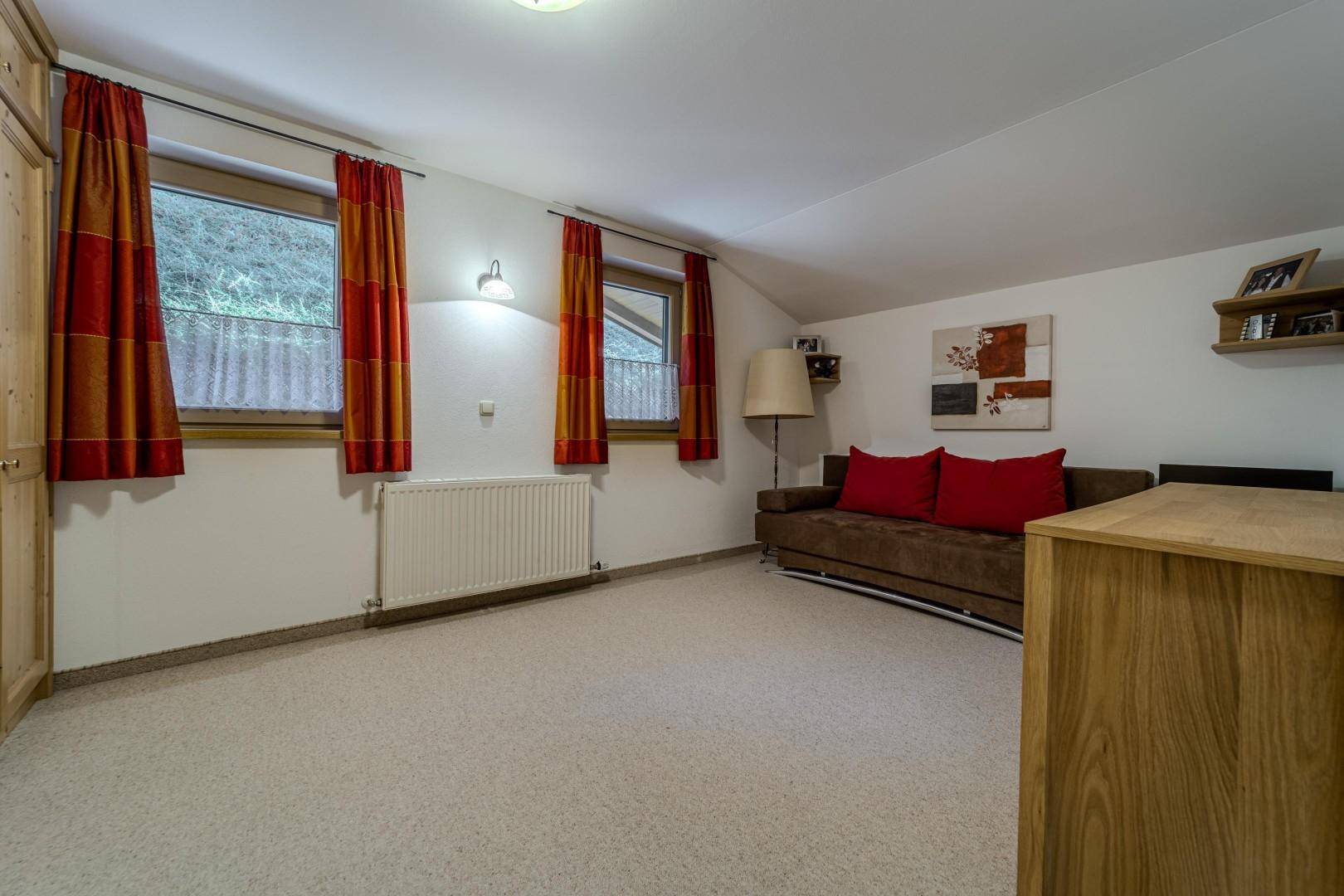 Gemütliches Zimmer mit Sofa, Teppichboden und Fenstern mit Blick ins Grüne.