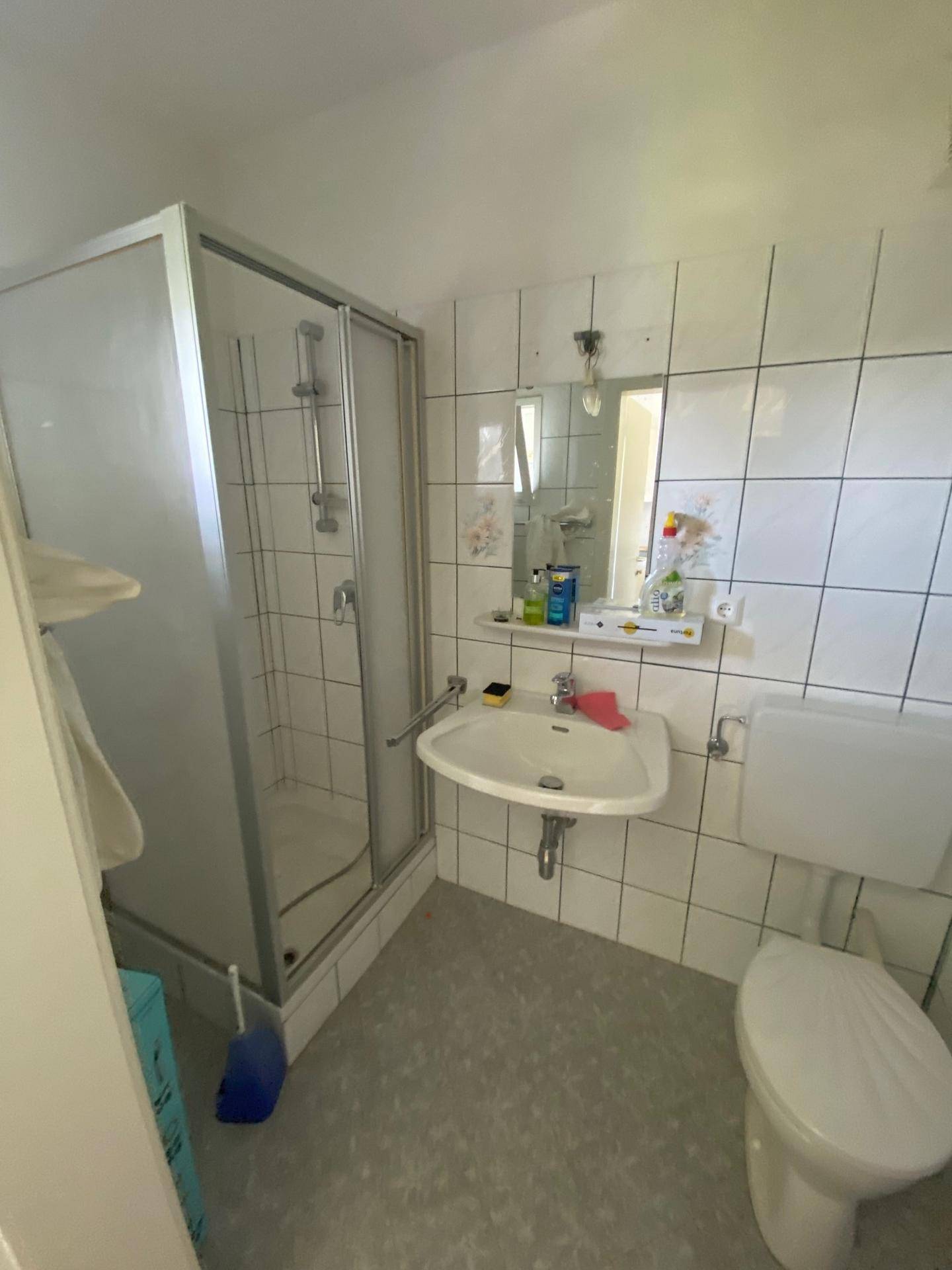 Älteres Badezimmer mit Duschkabine, Waschbecken und WC, gefliest in Weiß.