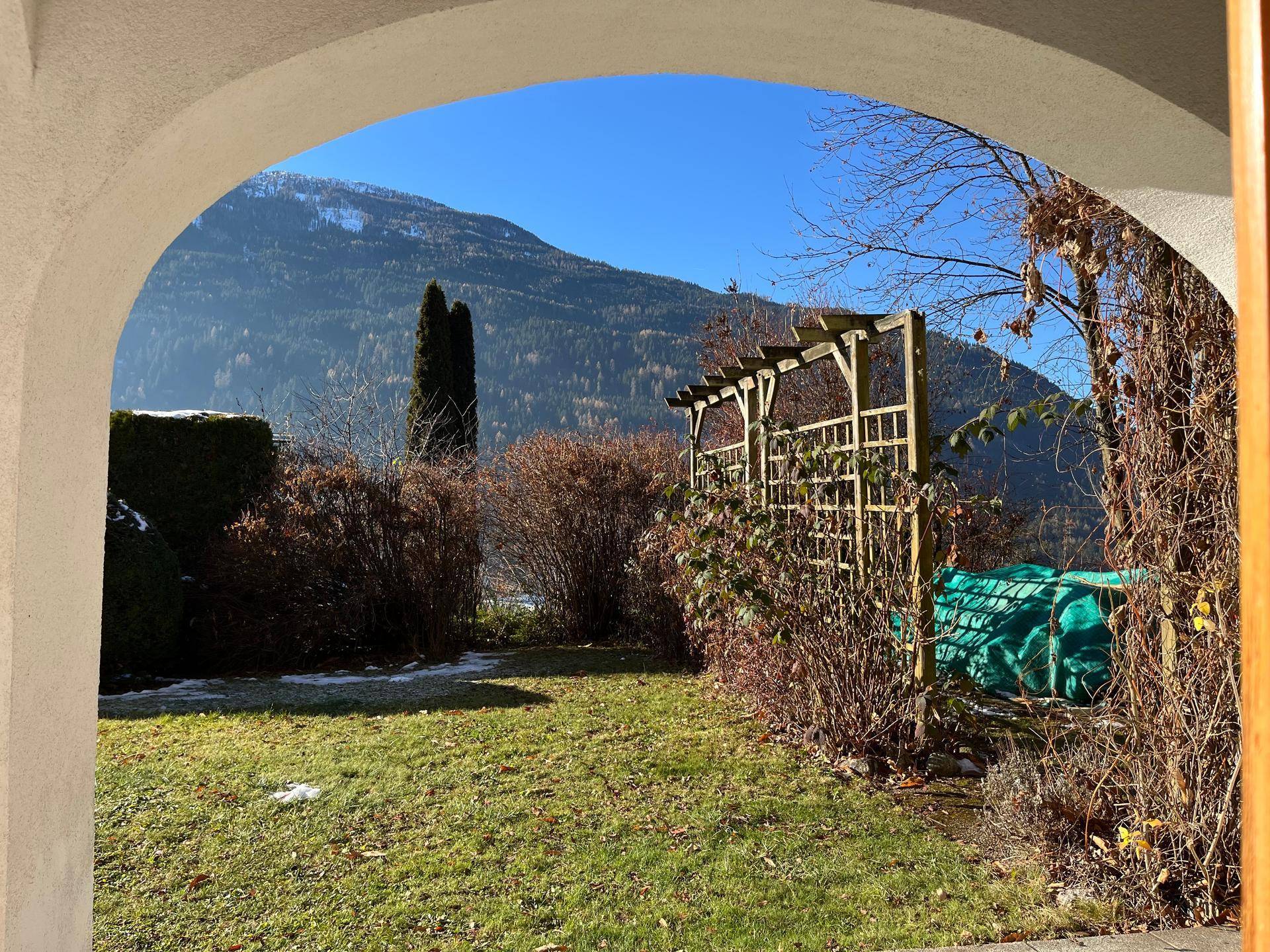 Blick durch einen Torbogen in einen gepflegten Garten mit Pergola und majestätischem Bergblick im Hintergrund.