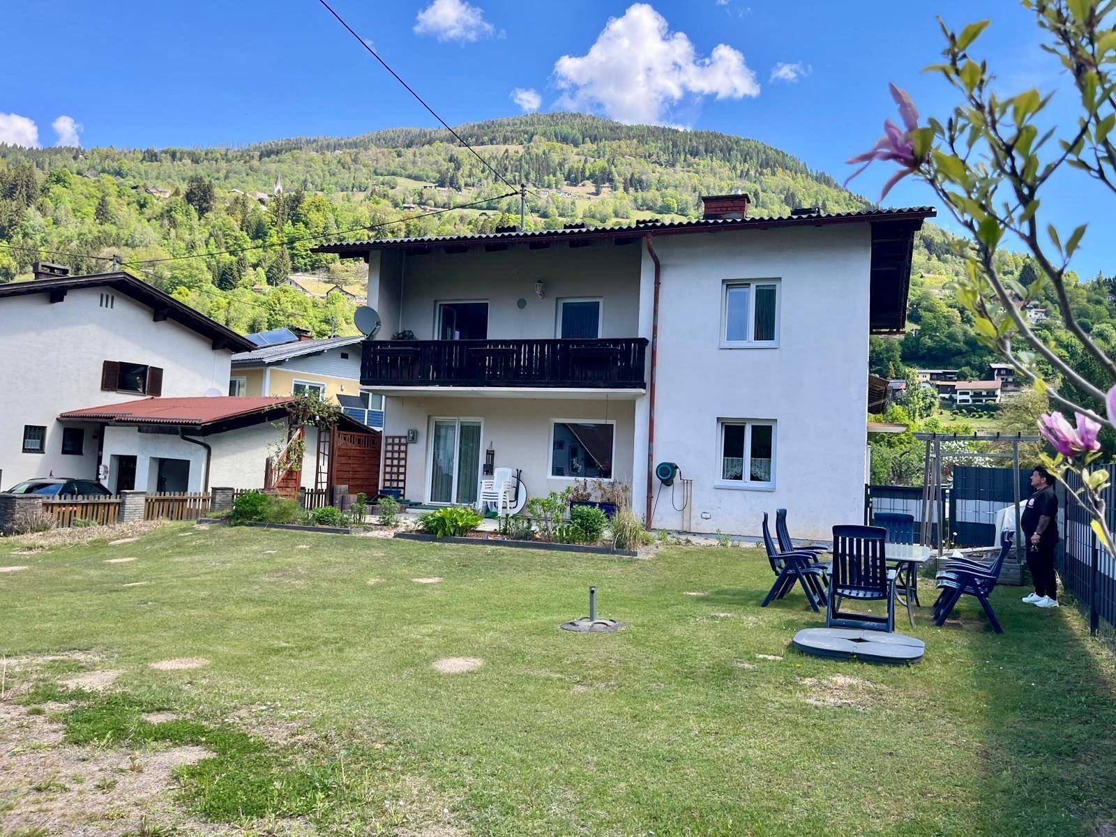 Hinteransicht des Hauses mit weitläufigem Rasen, Gartenmöbeln und Blick auf die umliegenden Berge.