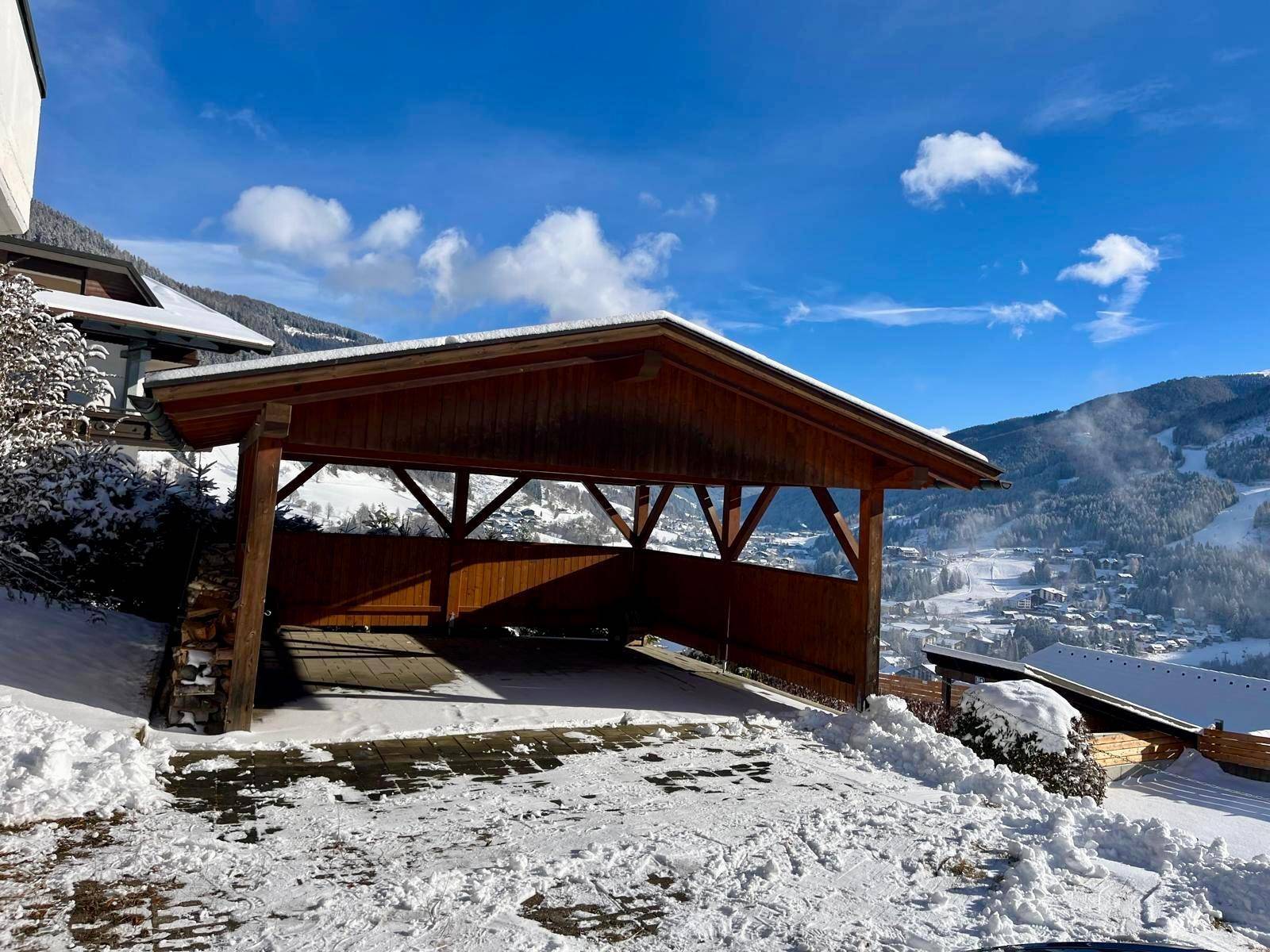 Holzcarport mit schneebedeckter Auffahrt und Blick auf die winterliche Berglandschaft.