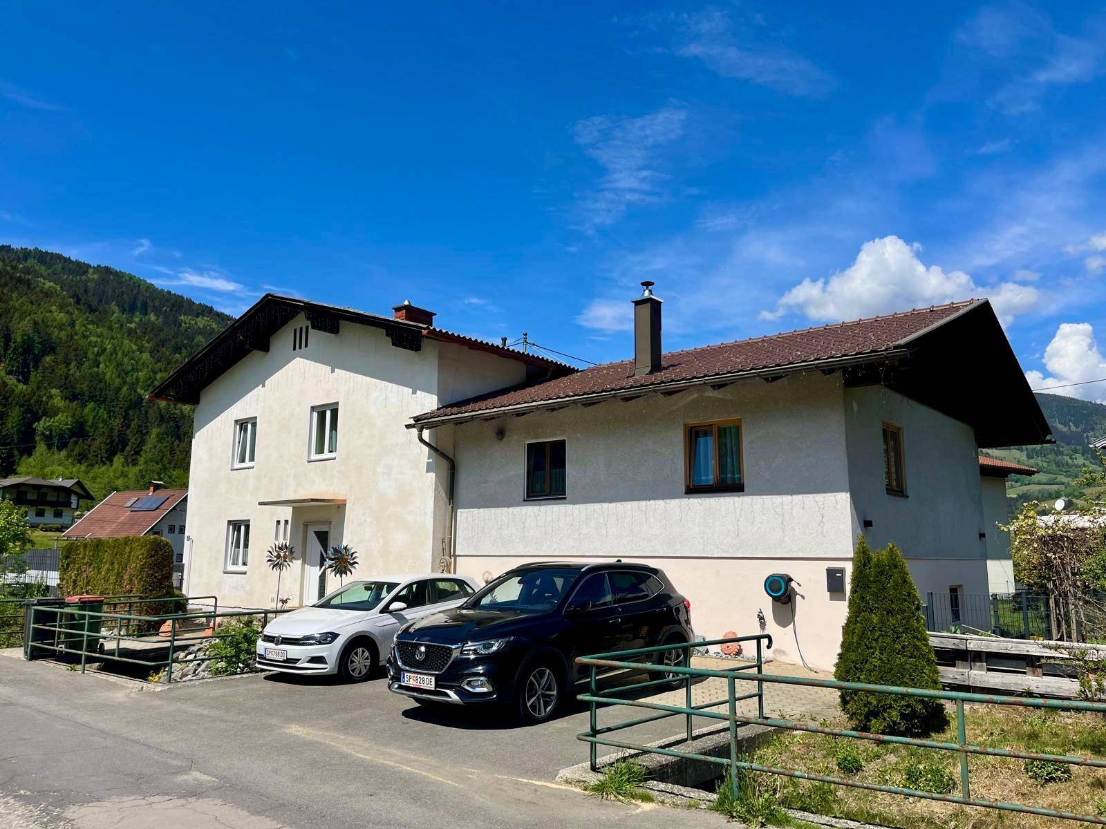 Vorderansicht des Hauses mit Einfahrt und geparkten Autos vor einer Bergkulisse.