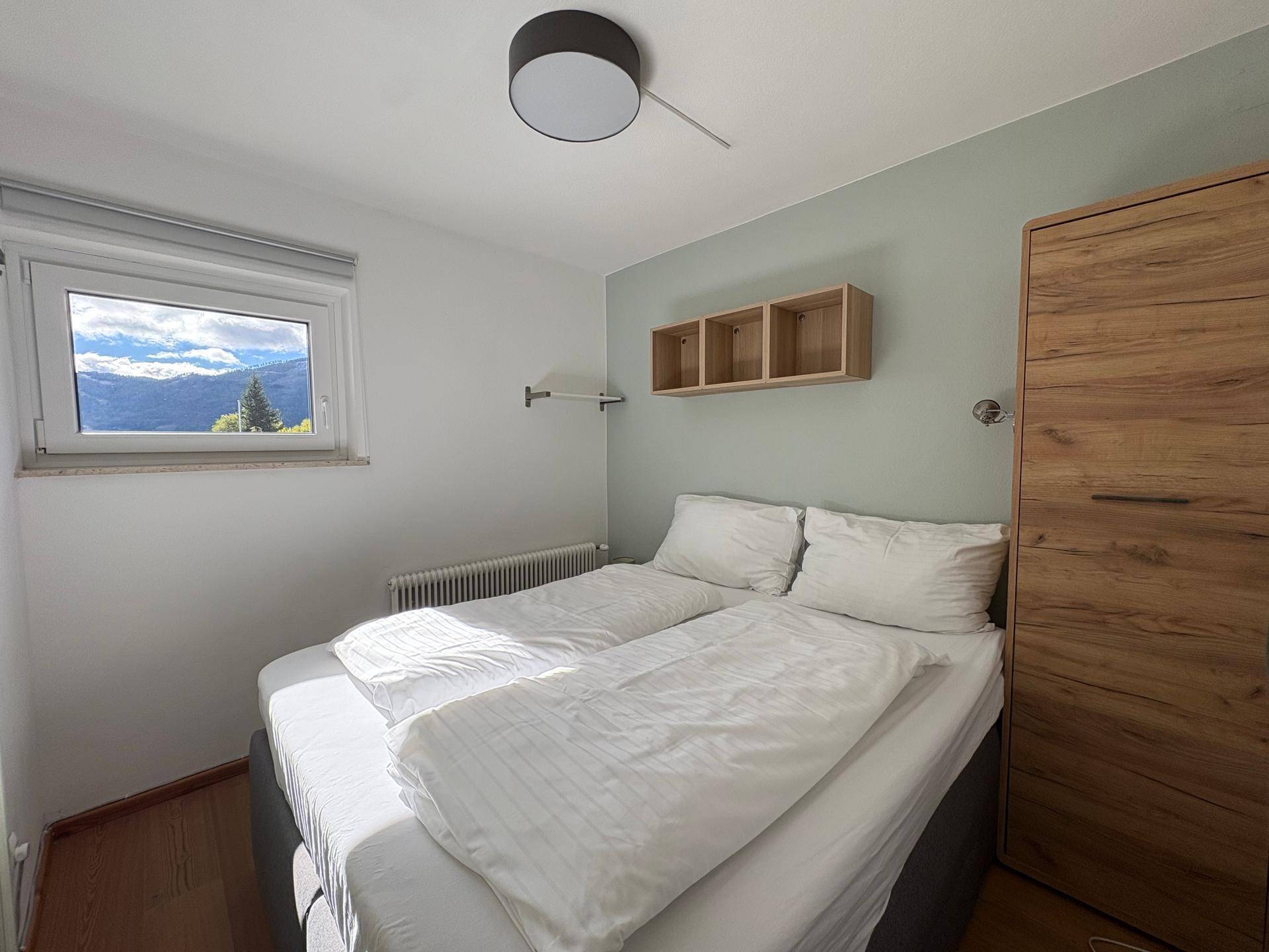 Schlafzimmer mit Doppelbett, Holzschrank und einem Fenster, das viel Tageslicht hereinlässt.