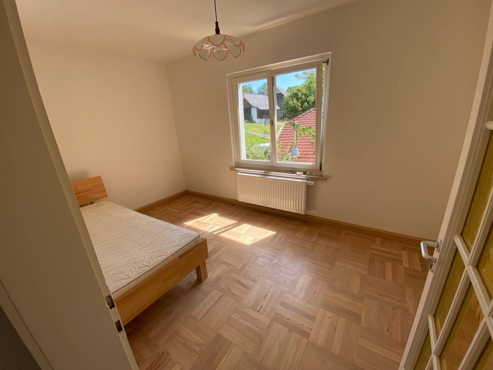 Helles Schlafzimmer mit Parkettboden und Fenster, das viel Tageslicht hereinlässt.