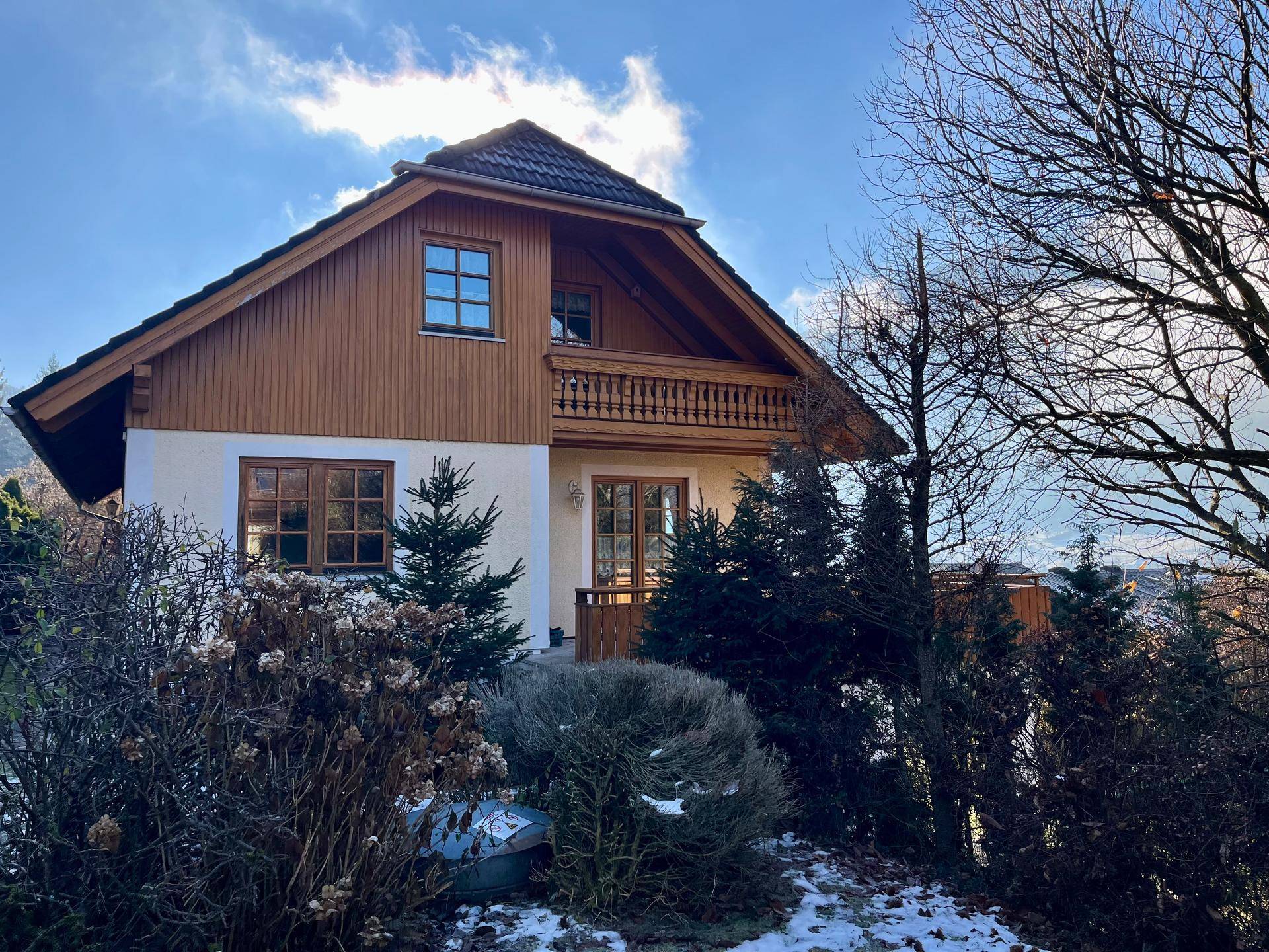 Charmantes Haus mit Holzfassade, Satteldach und einem kleinen Balkon, umgeben von winterlicher Vegetation.