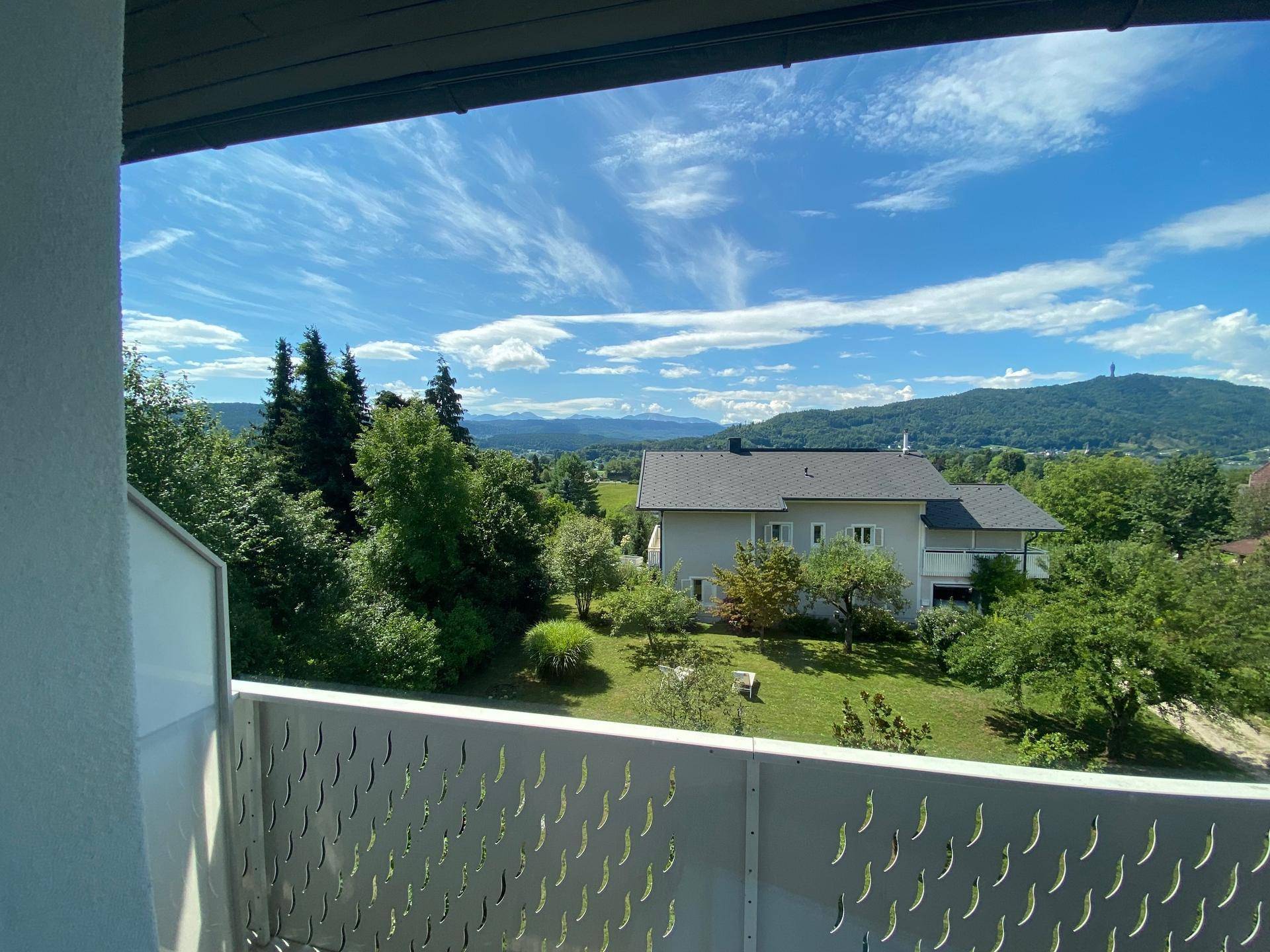 Blick vom Balkon auf die grüne Landschaft mit Bergen und einem Einfamilienhaus unter blauem Himmel.