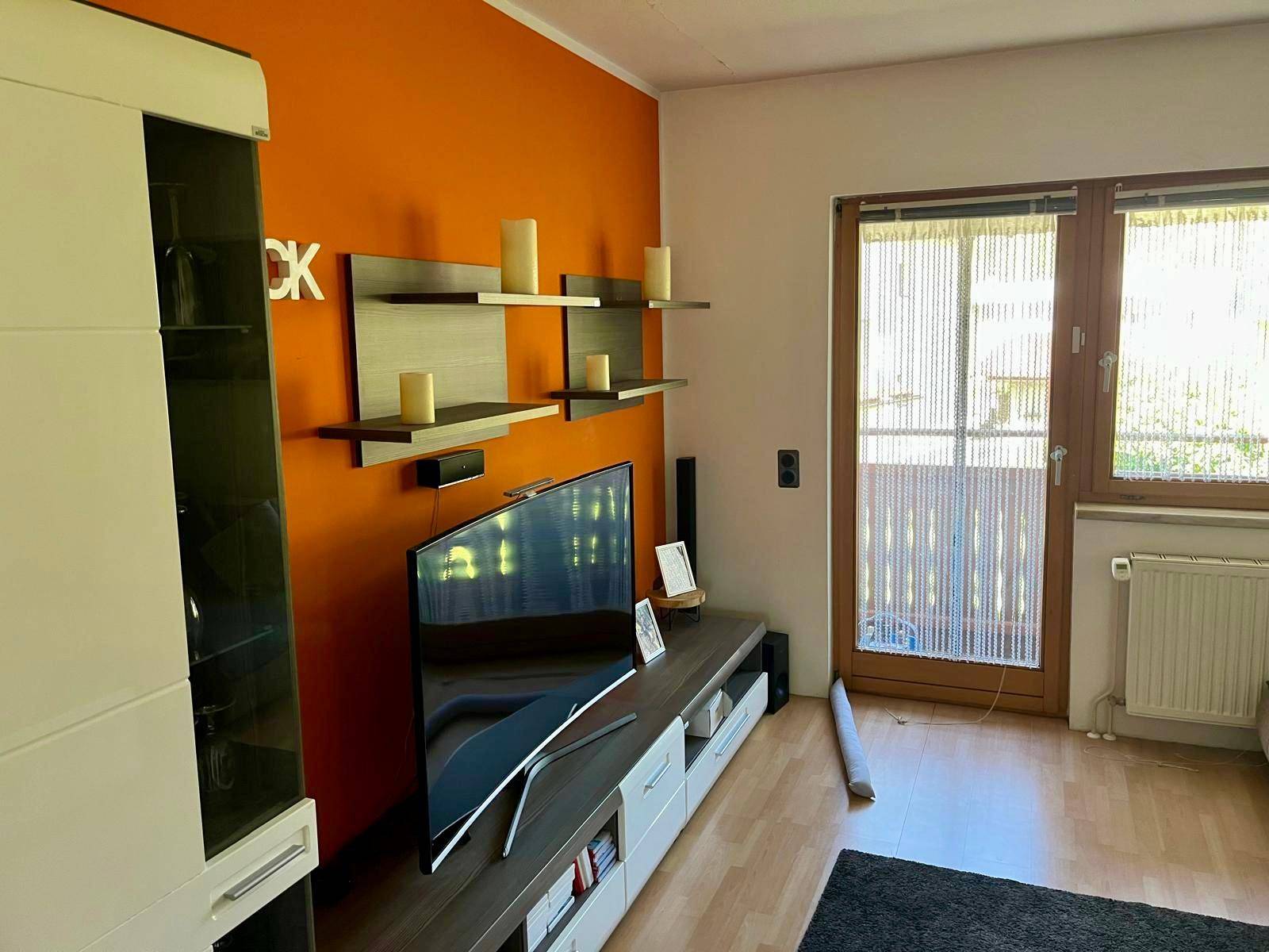 Wohnbereich mit orangefarbener Akzentwand, TV-Möbeln und Zugang zum Balkon.