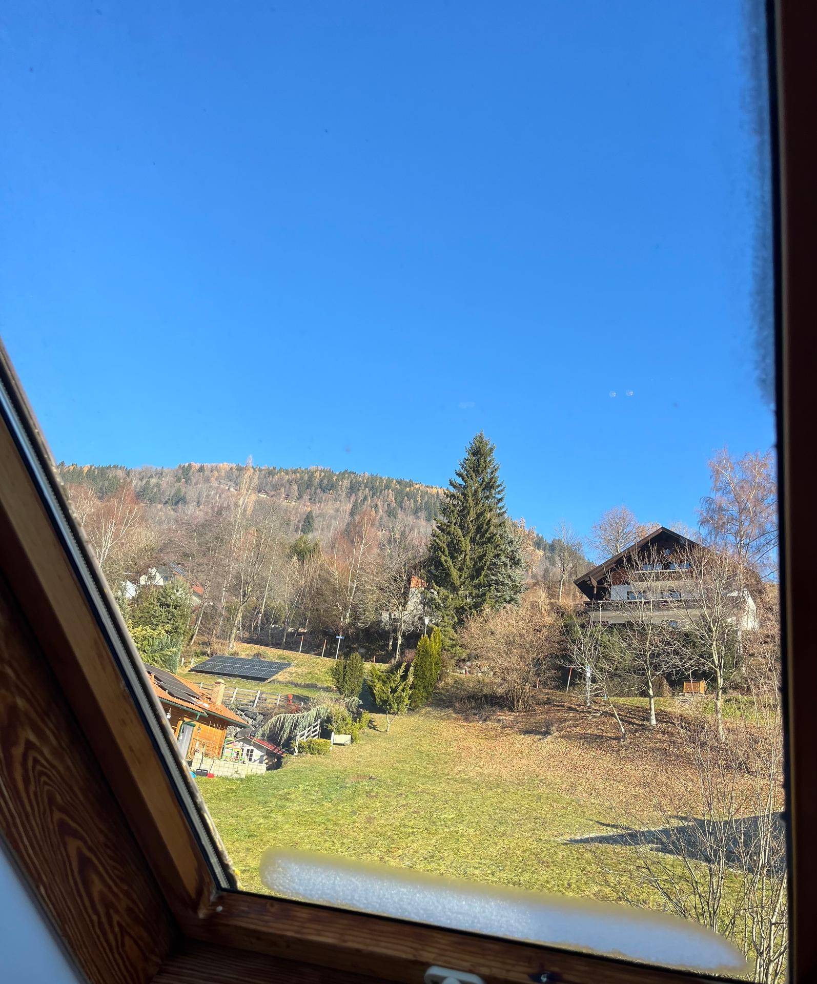 Blick aus einem Dachfenster auf eine grüne Wiese und bewaldete Hügel mit Häusern.