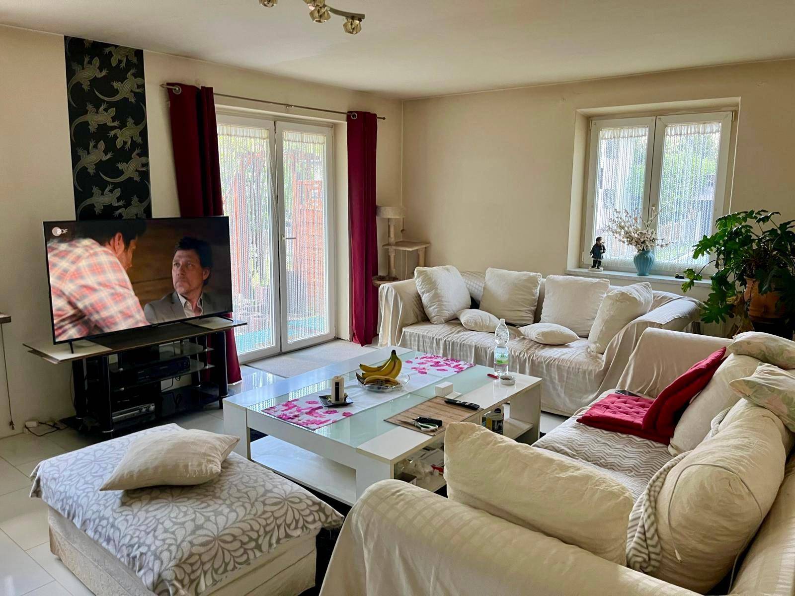 Helles Wohnzimmer mit großem Sofa, Fernseher und Zugang zu einem Außenbereich.