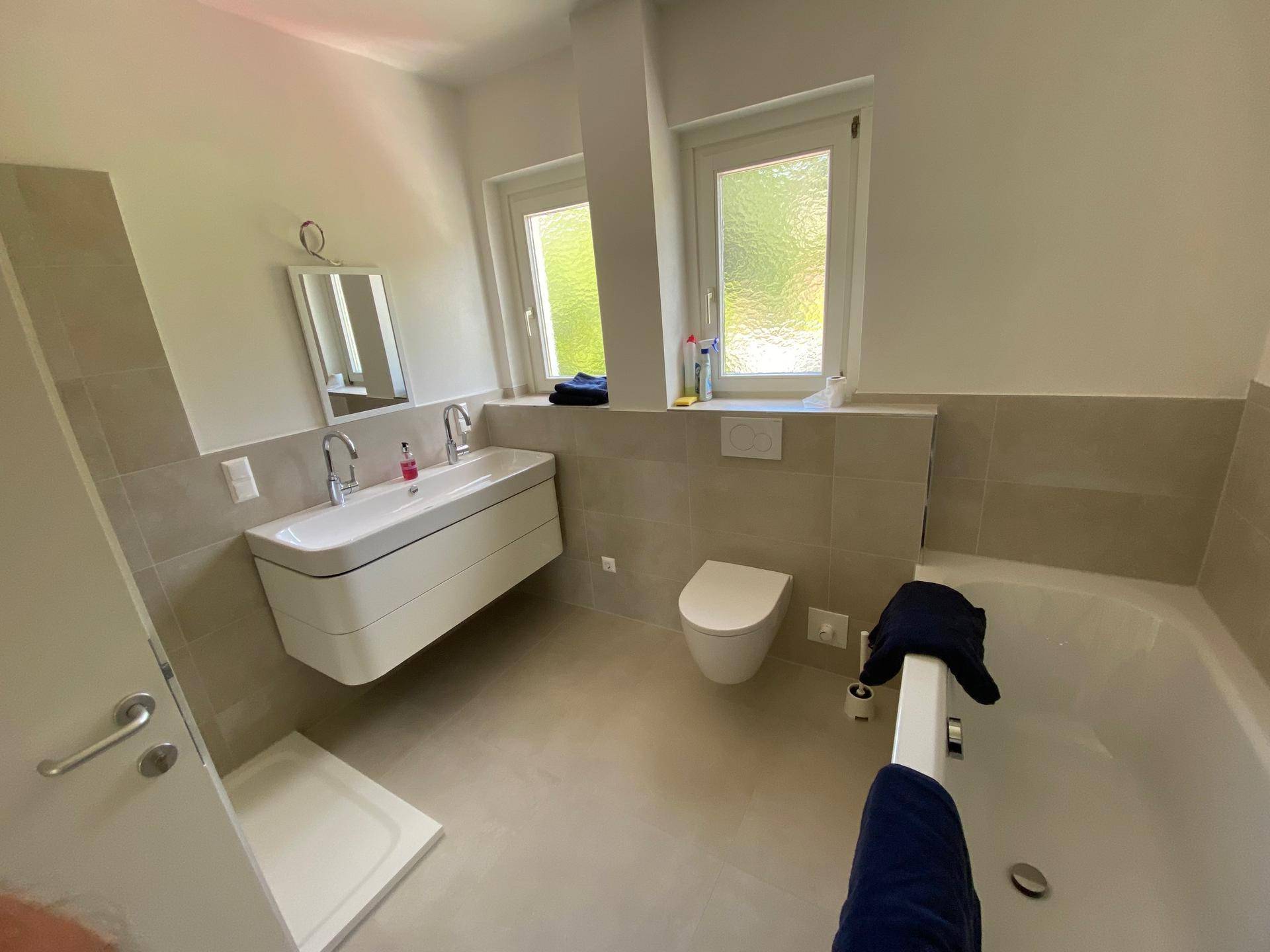 Modernes Badezimmer mit Badewanne, Dusche, Doppelwaschbecken und zwei Fenstern für Tageslicht.