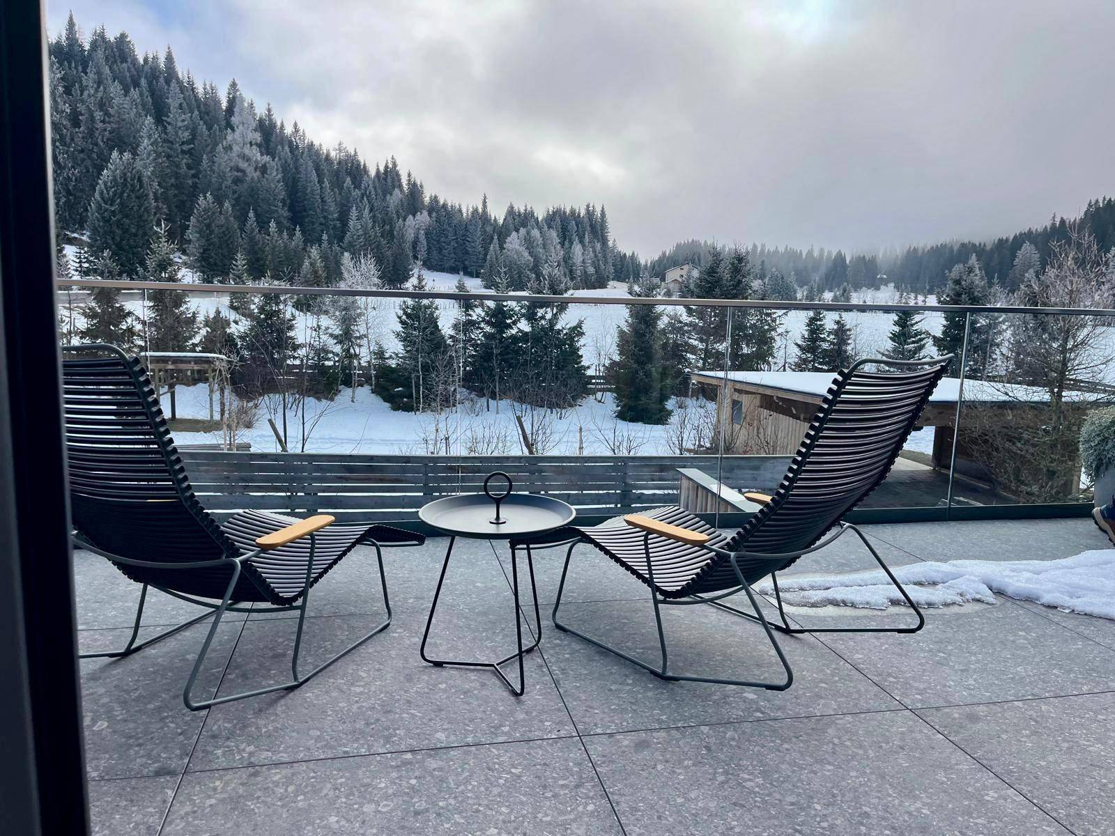 Terrasse mit bequemen Liegestühlen und weitem Blick auf die winterliche Berglandschaft und Wälder.