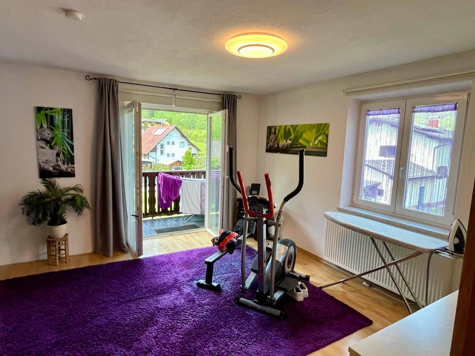 Geräumiges Schlafzimmer mit Fitnessgerät, lila Teppich und Balkonzugang.