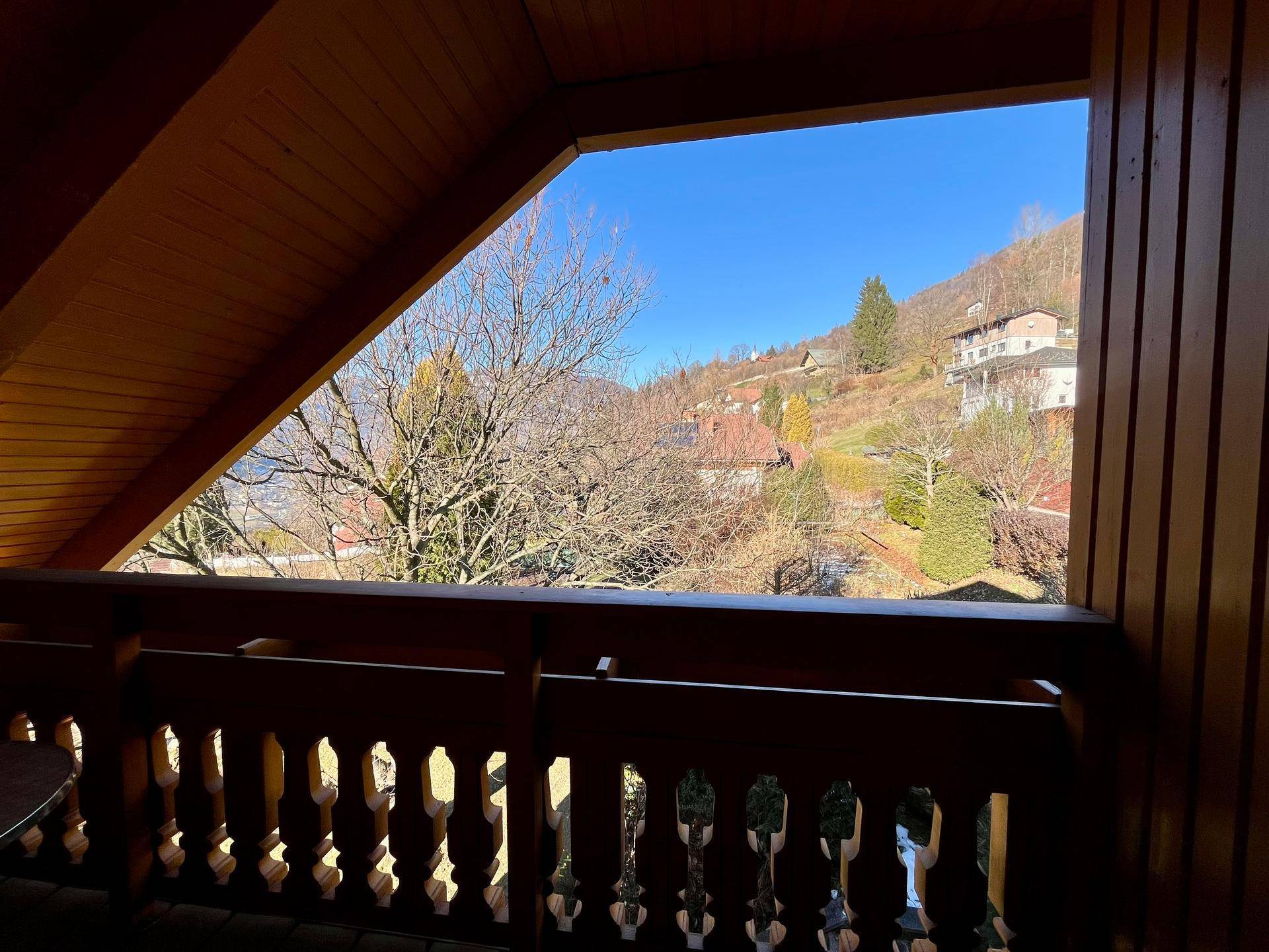 Sonnige Aussicht vom Holzbalkon auf die umliegende Hügellandschaft mit vereinzelten Häusern und Bäumen.