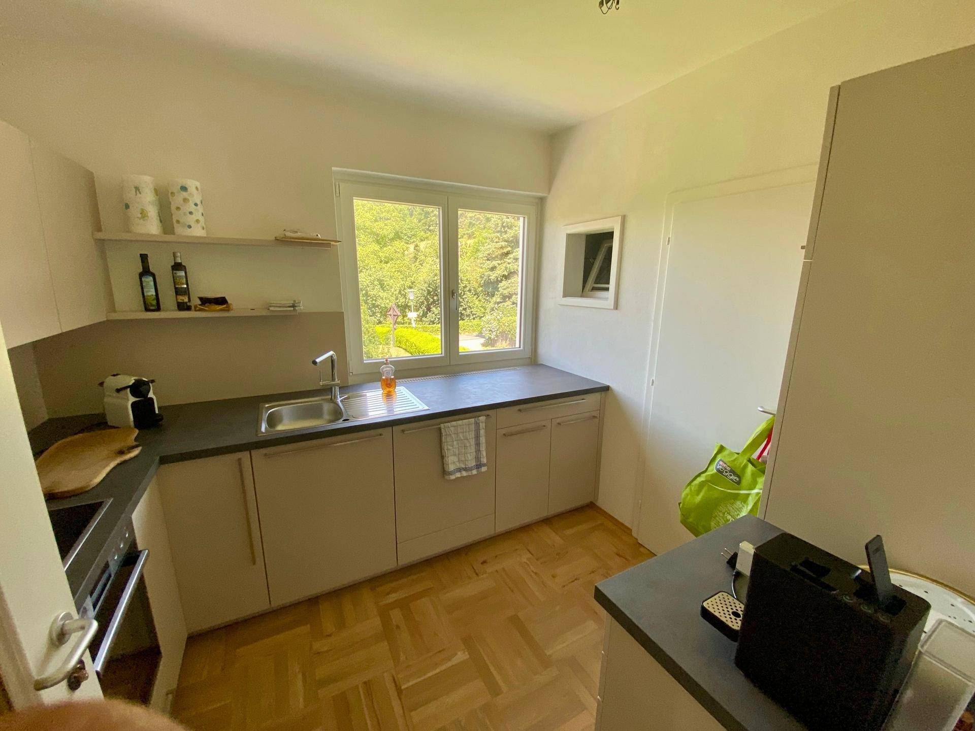 Funktionale Küche mit hellen Schränken, dunkler Arbeitsplatte und Blick ins Grüne durch das Fenster.