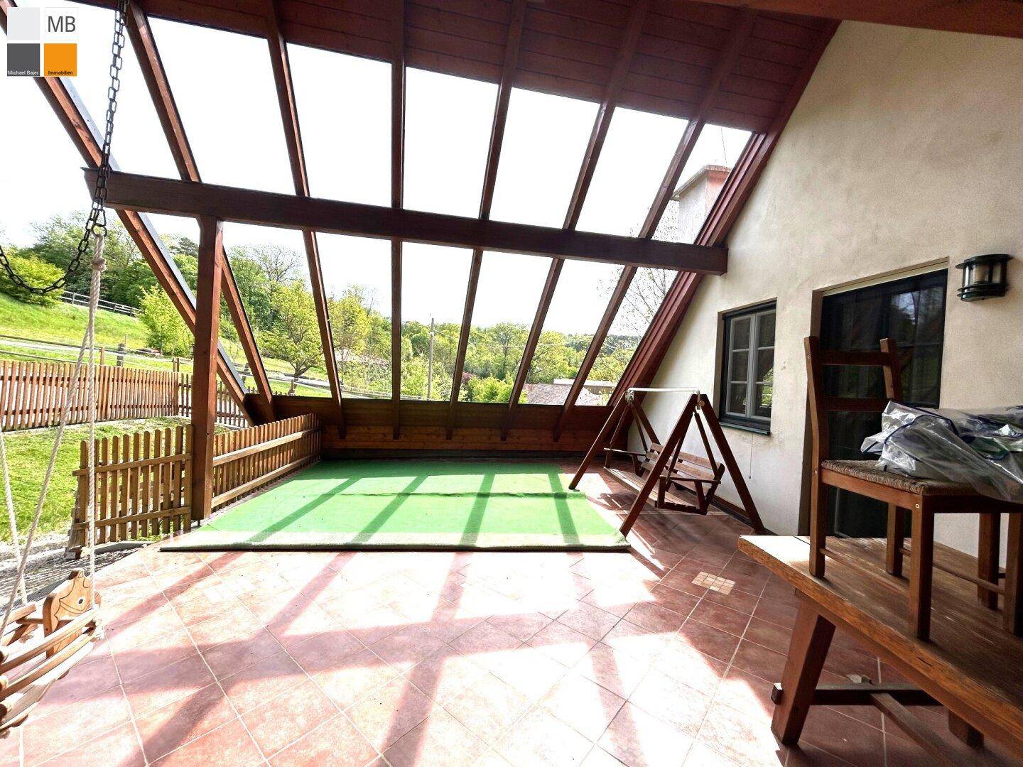 Überdachte Terrasse mit Fliesenboden, Schaukel und Blick ins Grüne.
