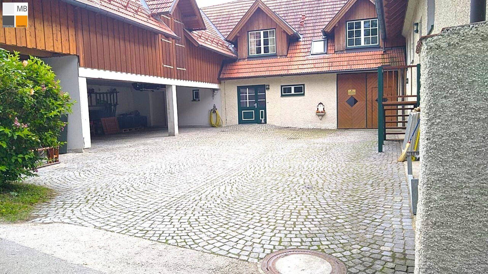 Traditionelles Gebäude mit gepflasterter Auffahrt und integrierter Garage, umgeben von Grün.