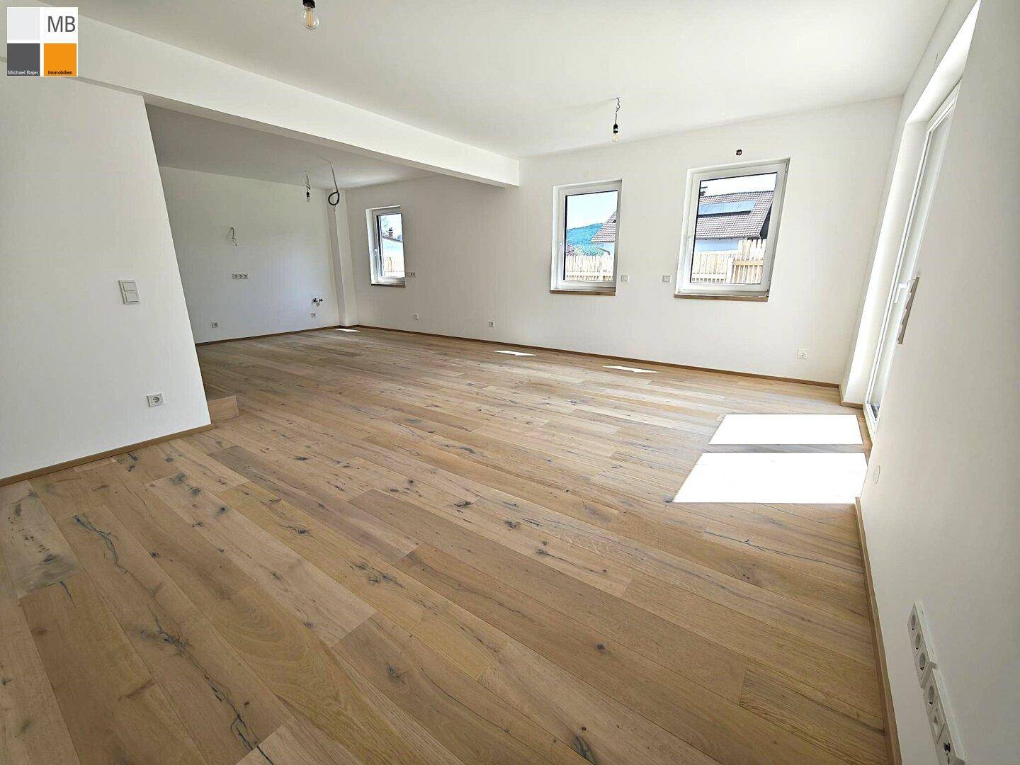 Geräumiges Wohnzimmer mit hellem Holzboden und mehreren Fenstern, die viel Tageslicht hereinlassen.