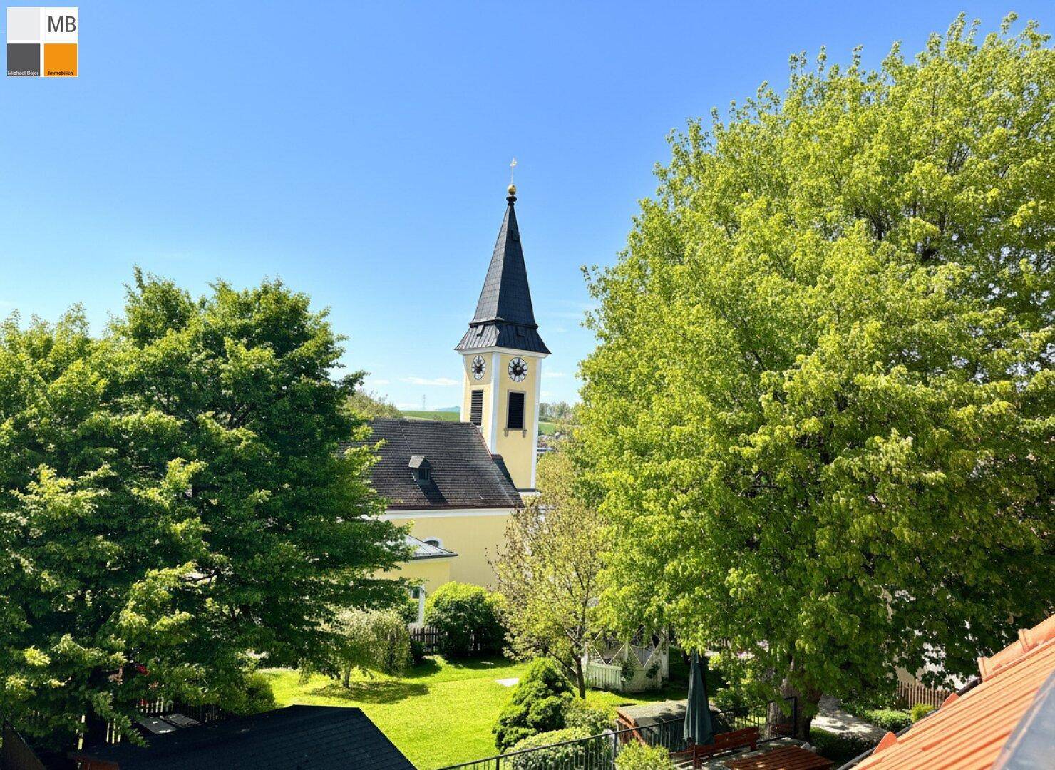Idyllischer Blick auf eine Kirche und grüne Bäume unter blauem Himmel.