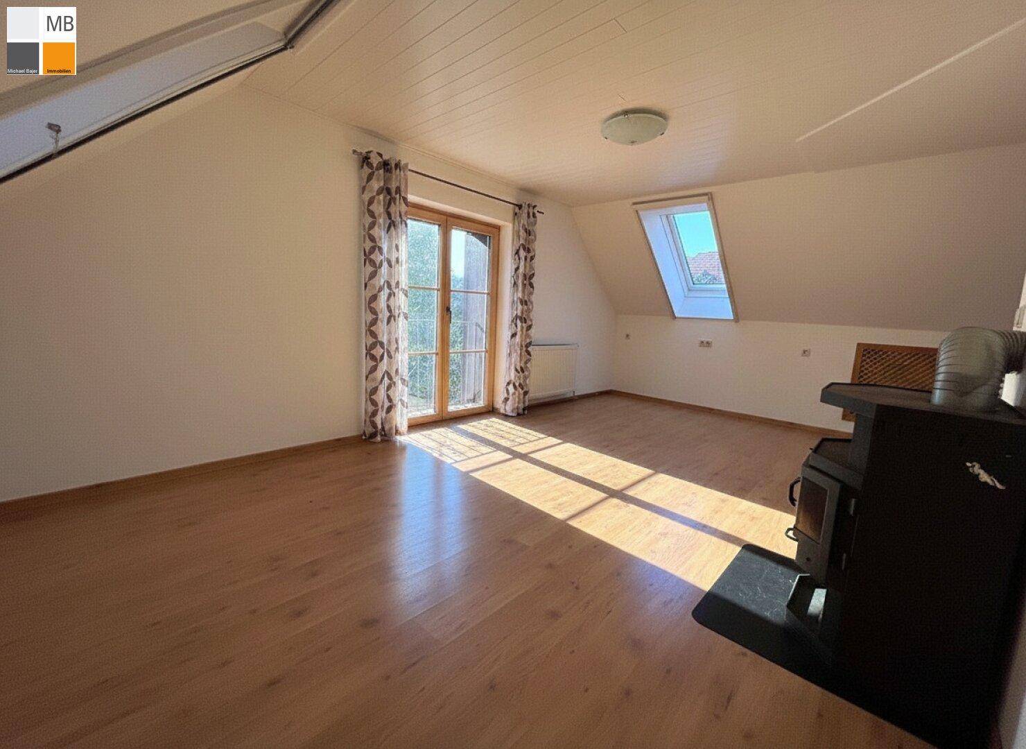 Sonniger Wohnraum im Dachgeschoss mit Holzboden, Kaminofen und großen Fenstern.