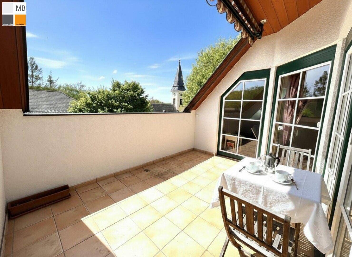 Sonniger Balkon mit Fliesenboden und Blick auf die umliegende Landschaft und Kirchturm.