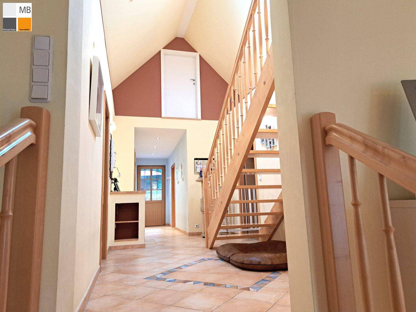 Geräumiger Eingangsbereich mit hoher Decke, Holztreppe und Fliesenboden.