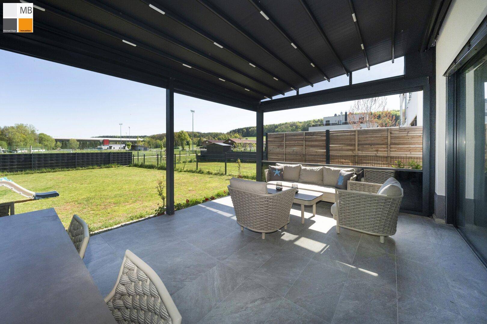 Geräumige überdachte Terrasse mit Loungemöbeln und Blick auf den Garten und Sportplatz.