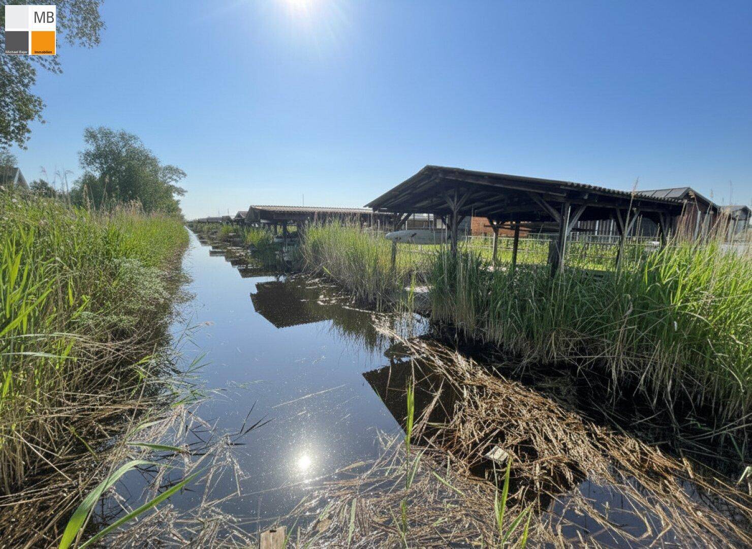 Schmaler Wasserweg zwischen Schilf und Holzhütten unter strahlendem Sonnenschein.