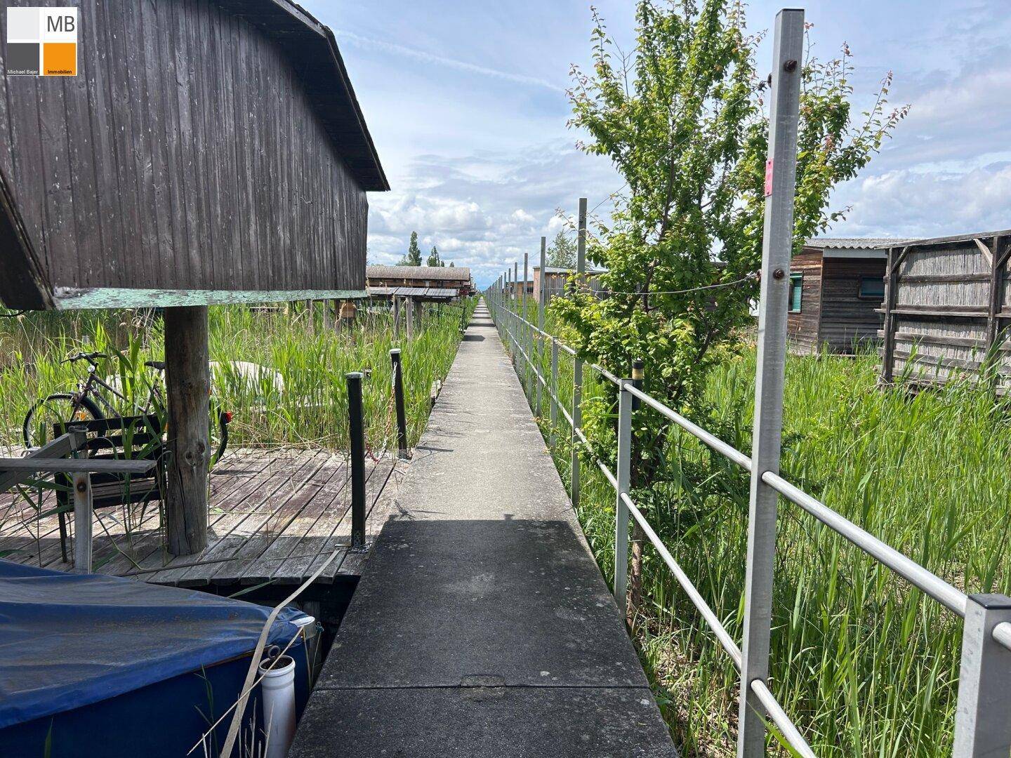 Betonweg zwischen Bootshäusern und grünen Ufern, führt entlang des Wassers.